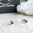 Birthstone Stud Earrings by Magnetic Mobility with blue Swarovski crystals. These earrings have a magnetic back and are ideal for fashionable pain relief, displayed on a reflective surface with Magnetic Mobility packaging.
