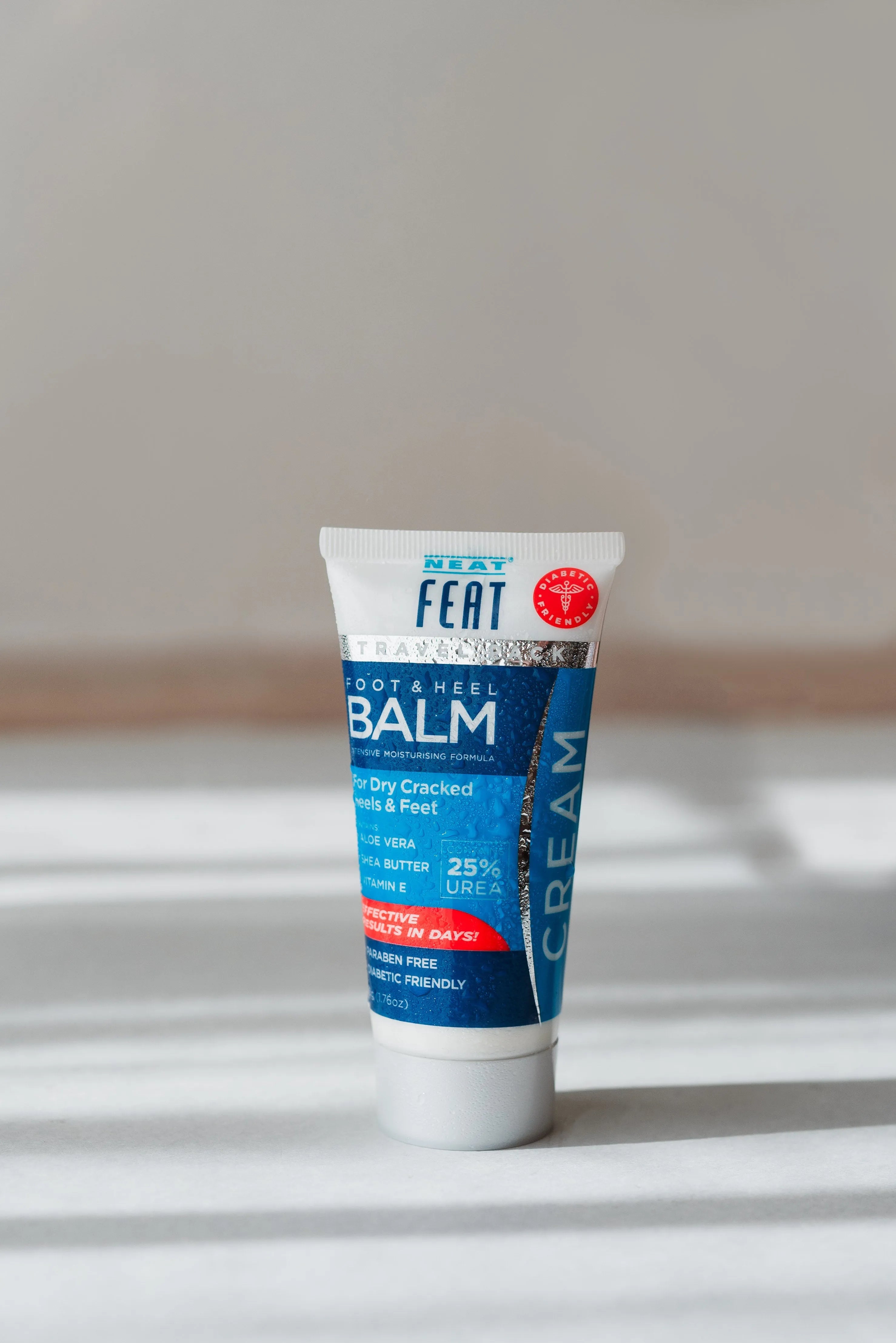 Front view of Neat Feat Foot & Heel Balm Travel Pack 50g tube, displayed on a grey surface with shadows and beads of water on the tube, featuring a grey cap and a blue label.