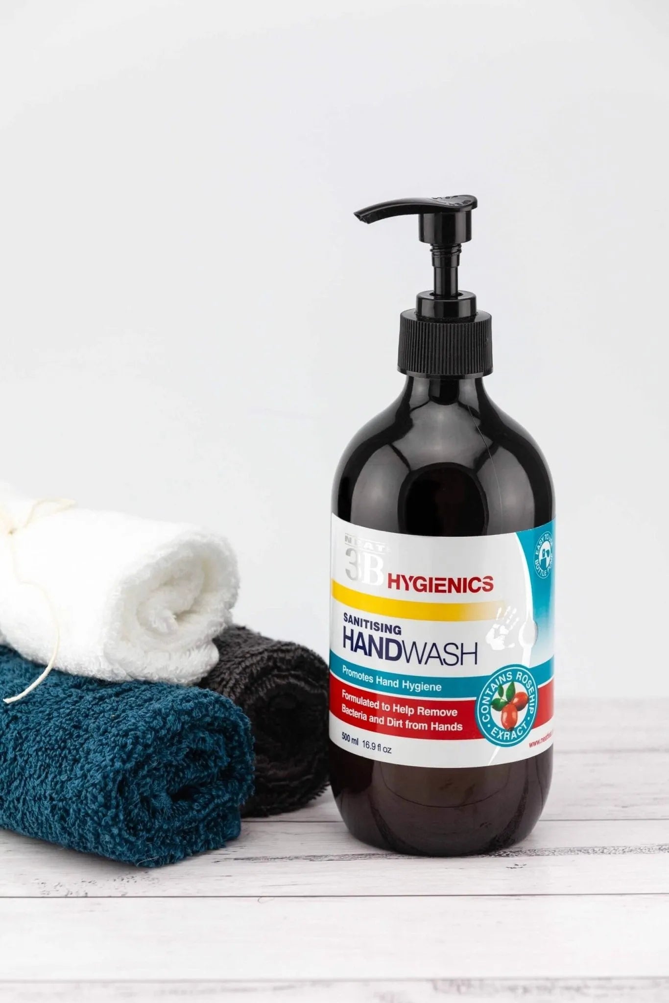 Neat Feat Hygienics Sanitising Handwash, front-on in a dark brown bottle with a black cap, featuring a white label with blue and red accents, displayed on a natural white table next to three small towels.